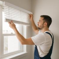 A man adjusts horizontal venetian blinds on a clear window with a silver lock and white surroundings.